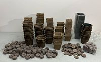 Full view of large quantity of compostable peat pots in round and square shapes, many compressed peat pellets, and tall black plastic plant pots.