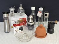 Full set of kitchen spice and seasoning tools arranged on table against black background. Includes various grinders, jars, garlic mincer in box, dressing and oil bottles, lemon squeezer, and sugar dispenser.
