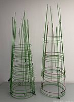 Photo of several green metal tomato cages standing upright, each constructed from multiple circular horizontal rings connected by vertical metal rods, designed to support tomato plants.