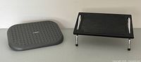 Overview of both under desk foot rests: black adjustable angled foot rest and grey textured foot rest side by side.