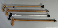 Four walking canes laid on flat surface: three wooden canes with natural oak finish, two identical 32 inch canes, one with metal table hanger; one light metal aluminum adjustable cane with black handle.