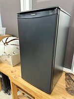 Front and side view of the black Danby compact refrigerator placed on a wooden surface against a wall and window background showing the door and overall unit shape.