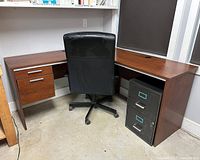 L-shaped wooden office desk with attached 2-drawer unit, black office chair in front, and black metal file cabinet on the right side