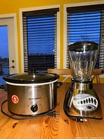 Side view of Hamilton Beach slow cooker and Oster blender showing full stainless steel surfaces, control knob and buttons, plugs visible