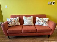 Front view of the sofa with three decorative cushions: two floral embroidered and one white textured cushion in the center. Sofa is pinkish-orange fabric with black legs.