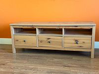 Front view of light brown pine TV bench with three drawers and three open compartments, showing wood grain and drawer knobs.