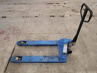 Side view showing entire pallet truck on warehouse concrete floor, blue paint with wear marks and steel forks with polyurethane wheels.