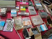 Box of assorted new screws, hooks, nails, and other handyman hardware items in original packaging on a red surface.