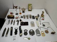Full view of all lot items displayed on a white surface, showing variety of bottle openers, lighters, bottle stoppers and various knick knacks including some decorative small figures and stands