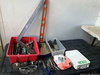 Overview of entire lot showing all included items arranged on a black surface and beside a wall. Red bin with screwdrivers, clear box with blades, safety glasses, first aid kits, scissors, level and carpenter square visible.