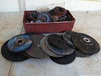 Plastic container holding various wire wheel brushes of different sizes and shapes with wire bristles, some rusted. Several cut off discs of multiple sizes are laid out in front of the container with visible wear and rust.