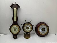 Three vintage barometers displayed side by side: wooden wall barometer with thermometer (glass missing), brass ship wheel barometer, and round wooden frame barometer.