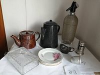 Overall view of vintage lot showing all items on table including wire mesh glass soda bottle, two tea pots, dessert plates, glass butter cover, and various small kitchenware items.