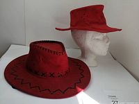 Front angle view of two red women’s hats on display: a leather cowboy hat with black leather stitching and a red felt hat on a white mannequin head.