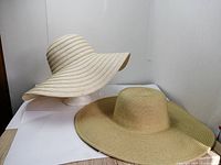 Two wide-brim wide summer hats, white LA Express hat on a white mannequin head and a natural straw hat placed on a white surface.