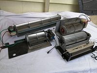 Five wood stove blower fans of varying sizes on a white surface with beige wall background.