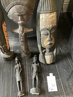 Five carved wooden African art pieces displayed on floor, including three statues and two wall art pieces with intricate carvings and patterns.
