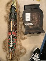 Photo showing a colorful elongated African mask with intricate painted patterns and natural fiber fringe beside a dark carved wooden box with a latching lid