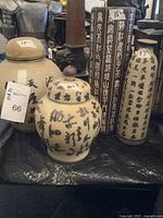 Photo showing three ceramic urns/vases with painted black calligraphy on cream/beige background. Includes a round lidded jar with a small figurine on lid, a tall rectangular vase with closely spaced calligraphy and a large urn marked '13"'.