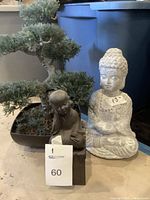 Photo showing two plaster Buddha statues (one large white and one smaller dark) and a plastic bonsai tree with green foliage in a black pot.