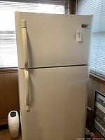Full front view of white Frigidaire refrigerator with top freezer, showing the two handles and standing near a window.