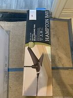 View of the boxed Hampton Bay ceiling fan showing front with fan image and product details.