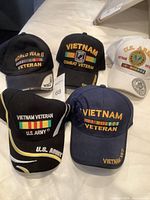 Photo shows five baseball caps with embroidered military veteran insignia and text, three black, one white, and one navy blue, representing Vietnam and World War II veterans.
