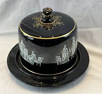 Overall front and side view of the black Pratware cheese dome and plate with visible white classical figures and gold trim.