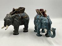 Photo of both cast iron elephant mechanical banks side by side on white background. One is blue, one is gray.