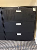 Single black business filing cabinet with three drawers, lock on the top drawer visible, note '3' label attached, shadow marks noticeable on top drawer.