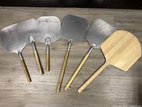 Six pizza paddles laid out side by side on a wooden floor, five metal with wooden handles, one fully wooden.