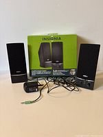 Front view of two black Insignia speakers with mesh covers and volume control on one speaker's base, along with power adapter and audio cable in front of original box.