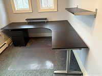 Black corner desk with adjustable legs and mini-shelf shown installed against wall under windows.