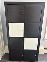 Front view of the IKEA cabinet showing 8 cubbies with doors; 6 black-brown, 2 white glossy doors with silver knobs.