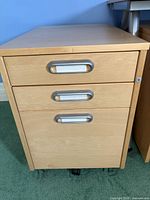 Front view of the IKEA light brown filing cabinet showing three drawers with metal handles and caster wheels below.