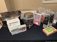 Full view of the lot including glass drink dispenser, multiple unopened packs of plastic glasses, napkins, paper straws, and a stack of napkins branded 'Fantastik'. Items are arranged on a black tabletop.