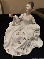Full view of the Royal Doulton figurine showing the seated woman in a white dress with gold floral decorations and holding a pink rose.