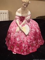 Photo of the seated lady figurine in a pink floral gown holding a fan, shown on black background.