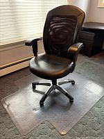 Front angle view of black pleather office chair on transparent plastic mat on carpeted floor next to window.