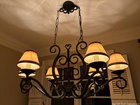 Full view of the black wrought iron chandelier showing all 6 lights with beige fabric shades lit, and scrolling wrought iron arms and frame attached to chain from ceiling mount.