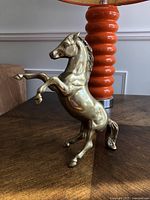 Photo of solid brass horse figurine standing on its hind legs with detailed mane and muscles, placed on wooden surface with an orange lamp in background.