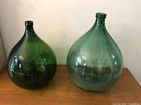 Two green glass demijohn bottles side by side on wooden surface with pale wall background.