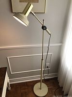 Full front view of the cream and brass colored adjustable floor lamp standing next to a window and wall paneling.