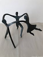 Steel sculpture of a dancing family with three abstract human figures interlinked by arms, matte black finish standing on three legs.