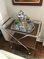Front angle view of the vintage chrome folding trolley bar cart with top wooden shelf holding a silver cocktail shaker and colored glasses on a silver tray.