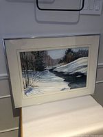 Framed watercolour painting showing a winter scene with snow and river, side angle.