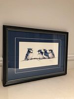 Framed watercolour painting showing the four baby blue jays perched on a branch with visible blue and white double matting and dark wood frame.