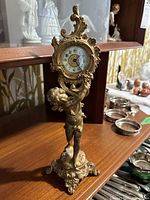 Full view of gilded cherub clock on wooden shelf showing base, figure, and clock face