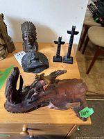 Photo showing two bronze figures, two resin totem poles in back, and a wood and resin souvenir plaque with carving of Indigenous craftsman on horseback on a table.
