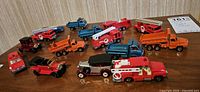 Fifteen vintage diecast toy cars and trucks arranged on a wood surface, showing variety of colors and types including fire trucks, dump trucks, and vintage cars.